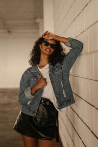 Chic young woman in denim jacket and sunglasses posing stylishly against a wall.