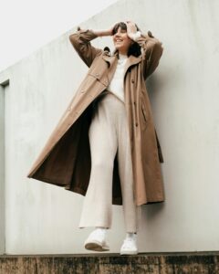 Low angle of full body of smiling female in trendy trench coat standing near building with light concrete wall and touching hair in daylight